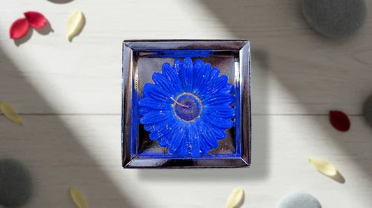Blue flower-shaped candle in square glass holder, decorative petals and stones on wooden surface