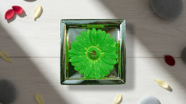 Green flower-shaped candle in a square glass holder on a light wooden surface with petals