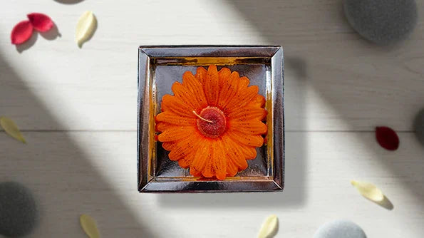 Orange flower-shaped candle in square glass holder on light wood background with petals and stones