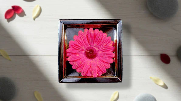 Pink flower-shaped candle in a square glass holder on a light wooden surface with petals