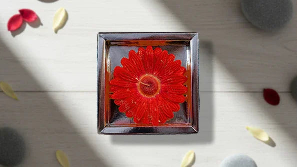 Red flower-shaped candle in square holder with petals and stones on a light surface
