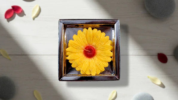 Yellow flower-shaped candle in a glass box on a light table with petals and stones