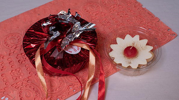 Flower-shaped candle with red center in clear dish beside red gift-wrapped package on pink cloth