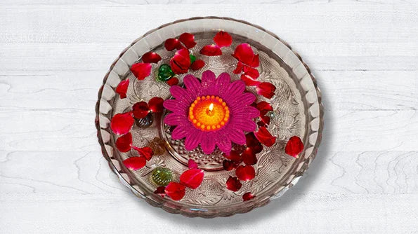 Pink flower-shaped candle floating in a glass bowl with rose petals on white background