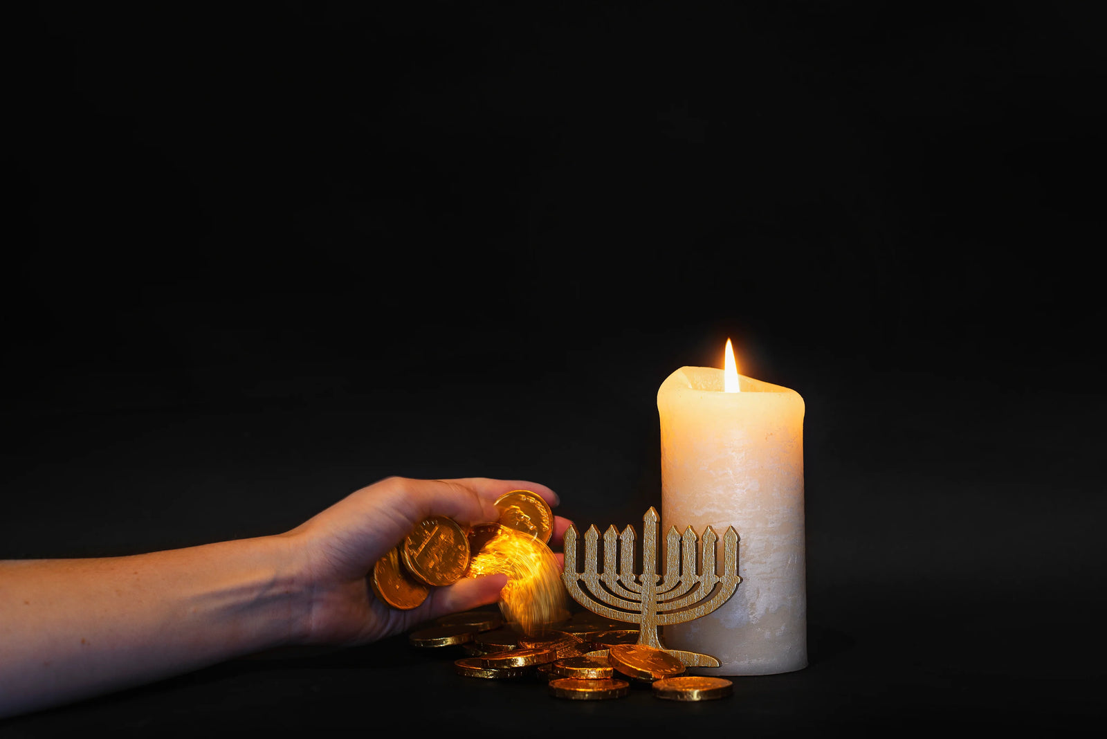 Gold Hanukkah gelt, menorah figurine, and lit candle on black background